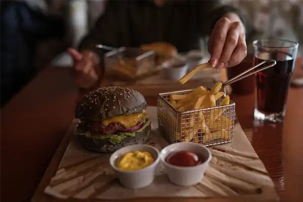 Tasty burger and sauce on wooden tray. Woman eating burger and chips in cafe. People and eating concept. Hamburger, french fries, ketchup, mustard on a wooden board. Toned image.